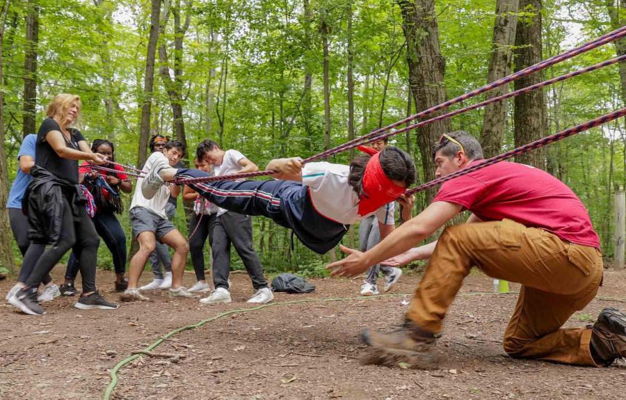 Milstein Program cohort in woods, pulling ropes to support a fellow student who is blindfolded and hovering horizontally above the ground