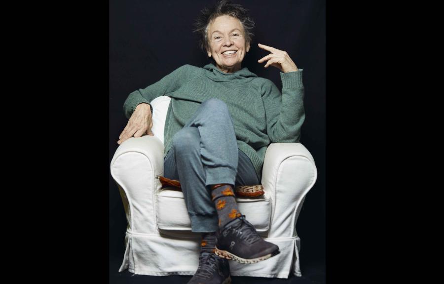 Laurie Anderson sitting in white chair against black background, smiling at camera